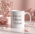 White mug with 'I Rise. I Heal. I Glow.' text on a pink floral background
